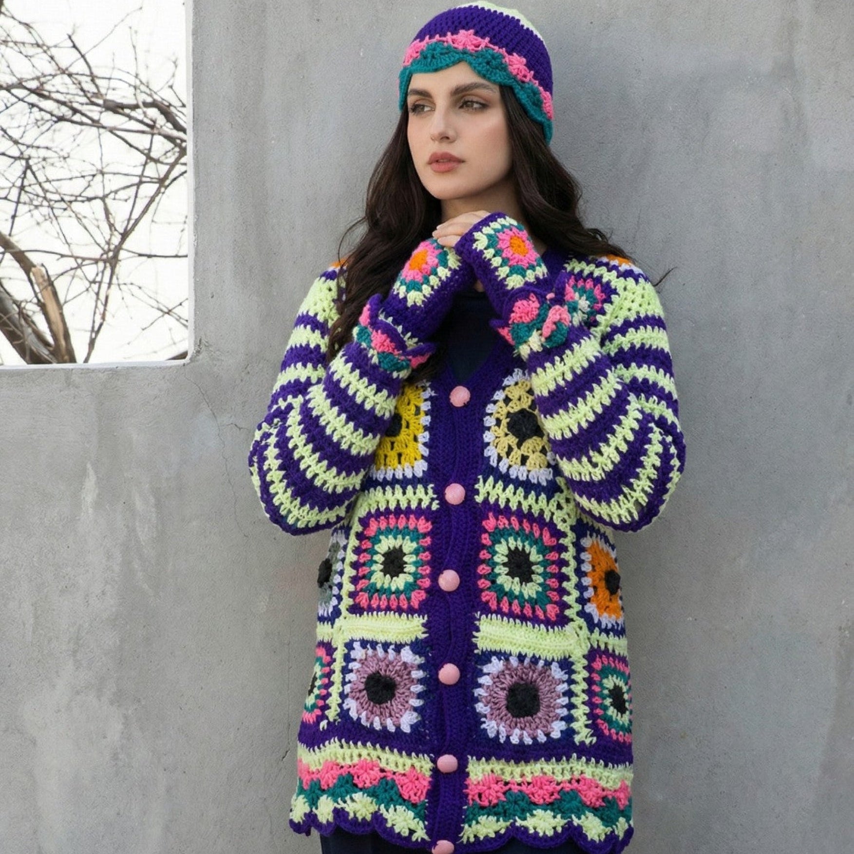 Woman wearing a colorful crocheted coat and hat against a gray wall.